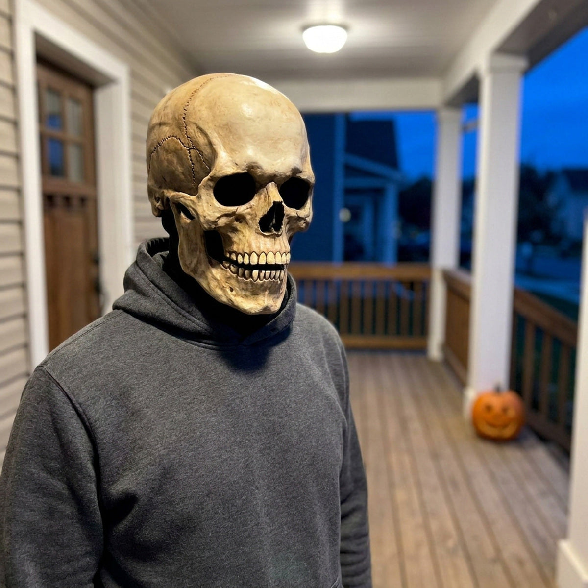 Adult wearing the aged bone skull mask on a lit front porch in the evening, a carved pumpkin further back