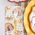 The Amber Harvest Table Setting by Sophistiplate features a white fork with a gold handle resting on a napkin adorned with colorful turkeys, accompanied by designer disposable plates and a patterned plate atop a plaid tablecloth, creating an inviting autumn tablescape.