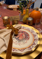 A Turkey & Gourds: Thanksgiving Table for 16 setting featuring a heavy duty paper plate with a turkey illustration, a beige napkin tied with a brown ribbon, and a place card labeled Nandinee, surrounded by gourds and candles on a red tablecloth.