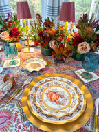 Turkey & Gourds: Thanksgiving Table for 16 set on a patterned tablecloth with gold chargers, teal glasses, and floral centerpieces with candles, featuring paper plates decorated with pumpkin illustrations.