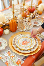 A hand arranges the Sophistiplate Turkey & Gourds Table Setting on an autumn table with Thanksgiving tableware—gold-rimmed plates, candles, glasses, mini pumpkins—plus a fall-patterned cloth and napkin.